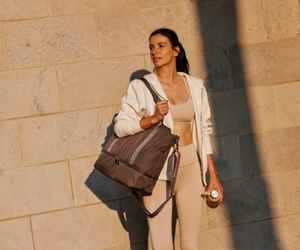 Woman with Lo & Sons Catalina Deluxe Tote, wearing athletic wear, holding a water bottle.